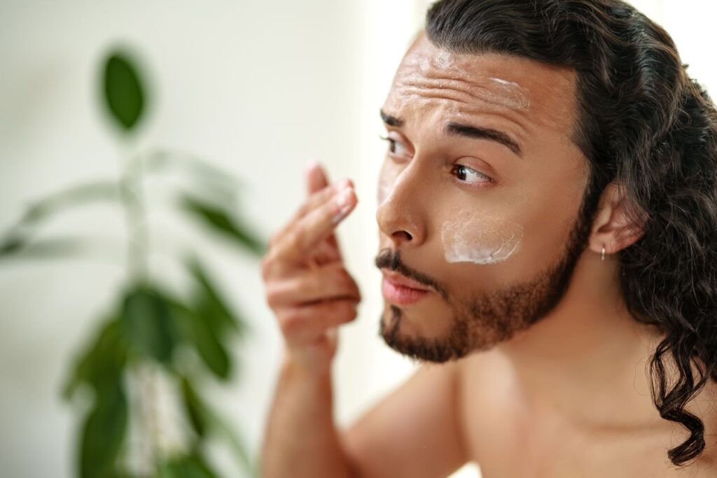 Mann mit langen Haaren und Bart trägt Gesichtscreme auf, um seine Haut nach einer Hydrafacial-Behandlung zu pflegen.